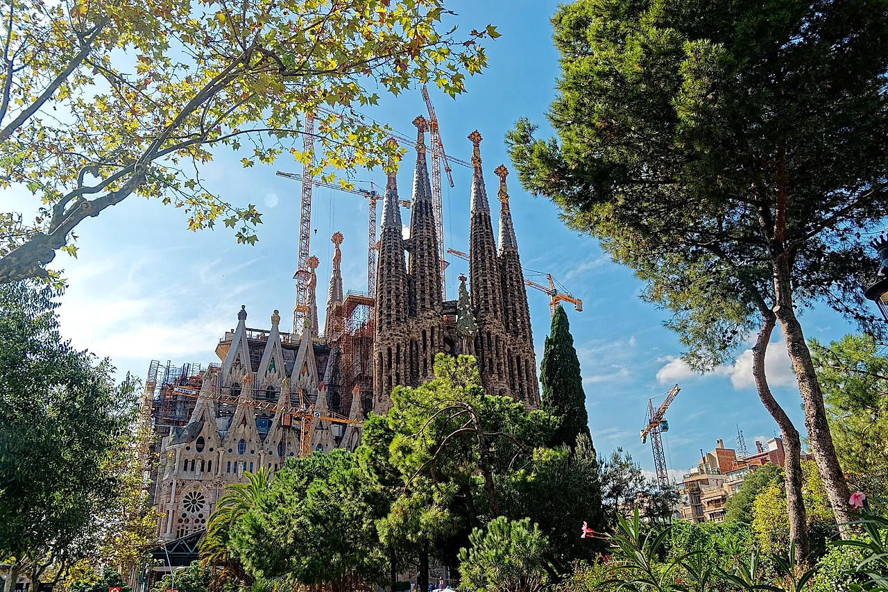 Le torri e la facciata della Natività viste tra gli alberi di Plaça de Gaudí, con le gru del cantiere sullo sfondo